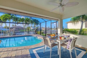A pool area with a table and chairs, a ceiling fan, and a view of palm trees and houses. - Villa Barefoot Cove (Cape Coral)