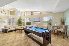 A pool table in a living room with a sofa and chairs, a dining table with chairs, and a window with blinds. - SeaBreeze Vista (Cape Coral)