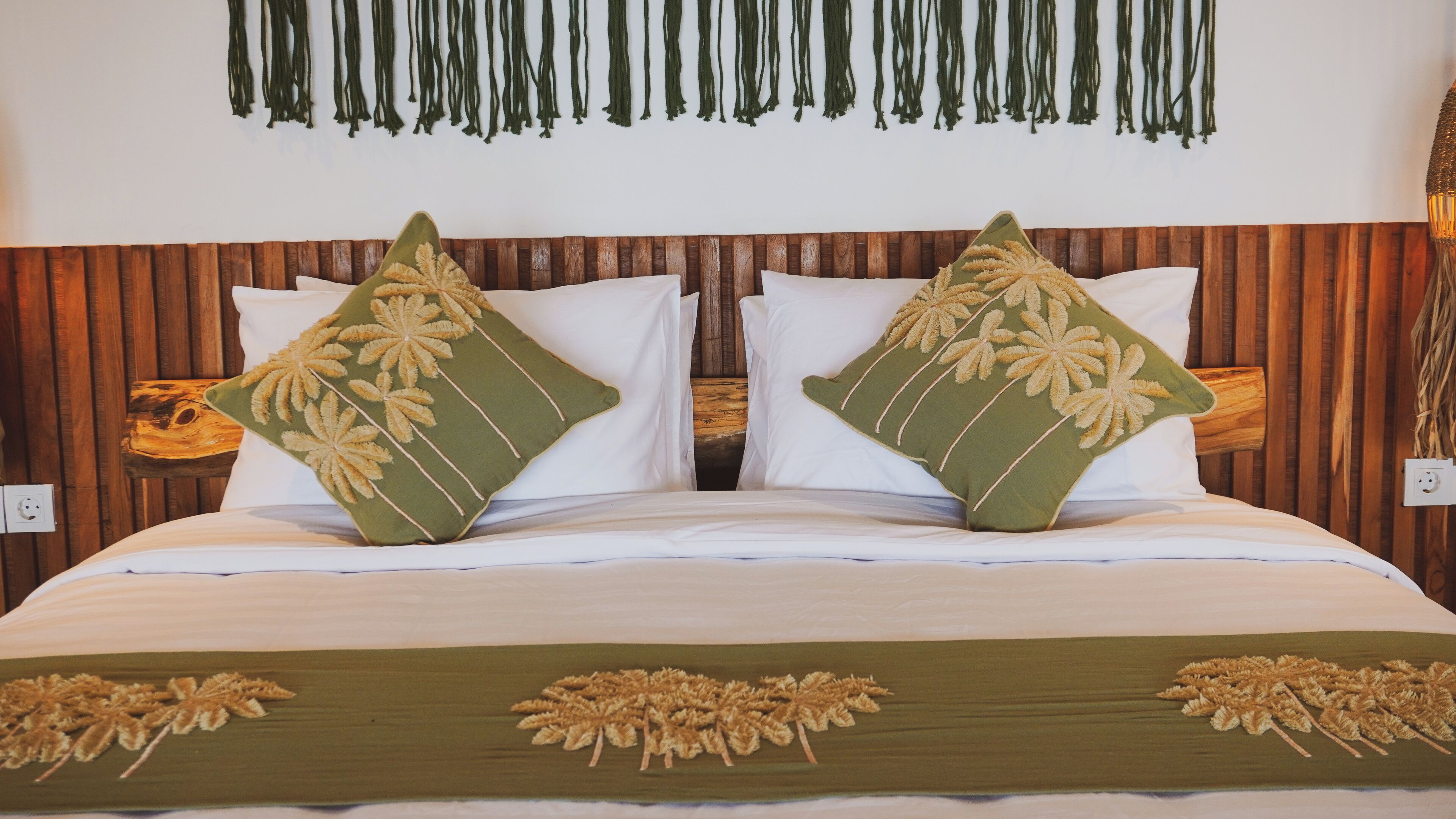 Una cama con dos almohadas verdes decoradas con hojas de palma, un cabecero de madera y una colcha blanca con diseños similares.