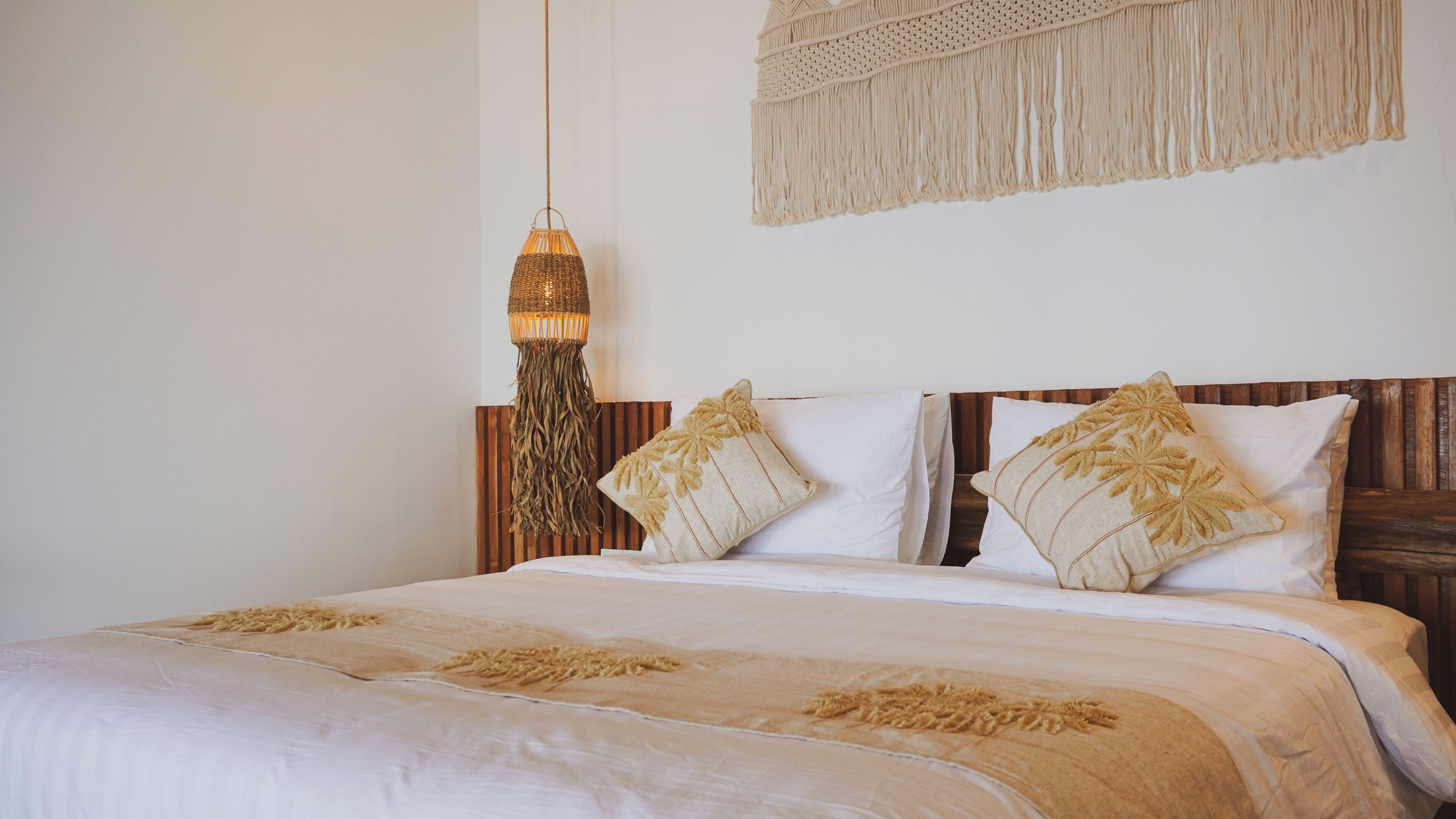 A neatly made bed with a beige and white color scheme, a hanging macrame wall hanging, and a woven pendant light.