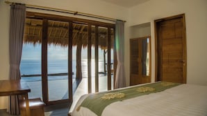 A bedroom with a large bed, a wooden desk, and a view of the ocean through a sliding glass door.