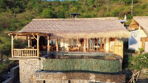 A thatched-roof building with a pool, surrounded by stone walls and a lush hillside.