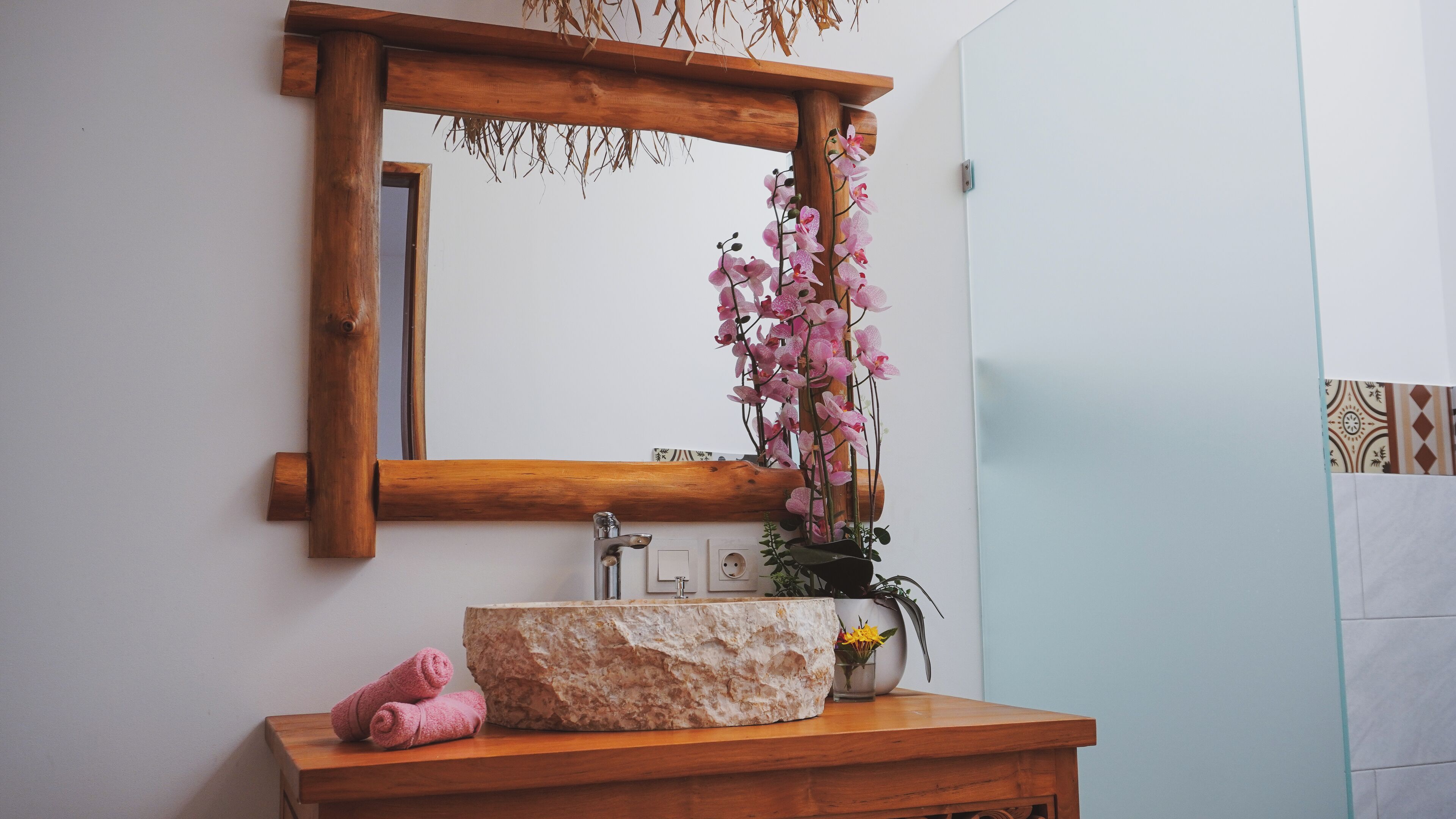 Une salle de bain avec un meuble-lavabo en bois, un vasque en pierre, un miroir, des serviettes roses et un vase contenant des orchidées roses.