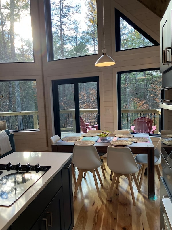 A modern kitchen with large windows, wooden flooring, and a dining area with chairs and a table set for four. - Modern Cabin 3bedroom 2bathroom- Whiteface Adirondacks (Jay)