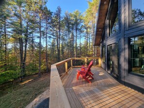 A wooden deck with a red chair, surrounded by tall trees and a modern building with large windows. - Modern Cabin 3bedroom 2bathroom- Whiteface Adirondacks (Jay)