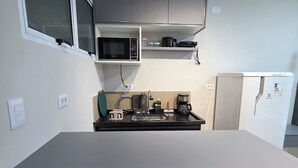 Et lille køkken med en hvid bordplade, to sorte barstole, en mikrobølgeovn, kaffemaskine og et køleskab.