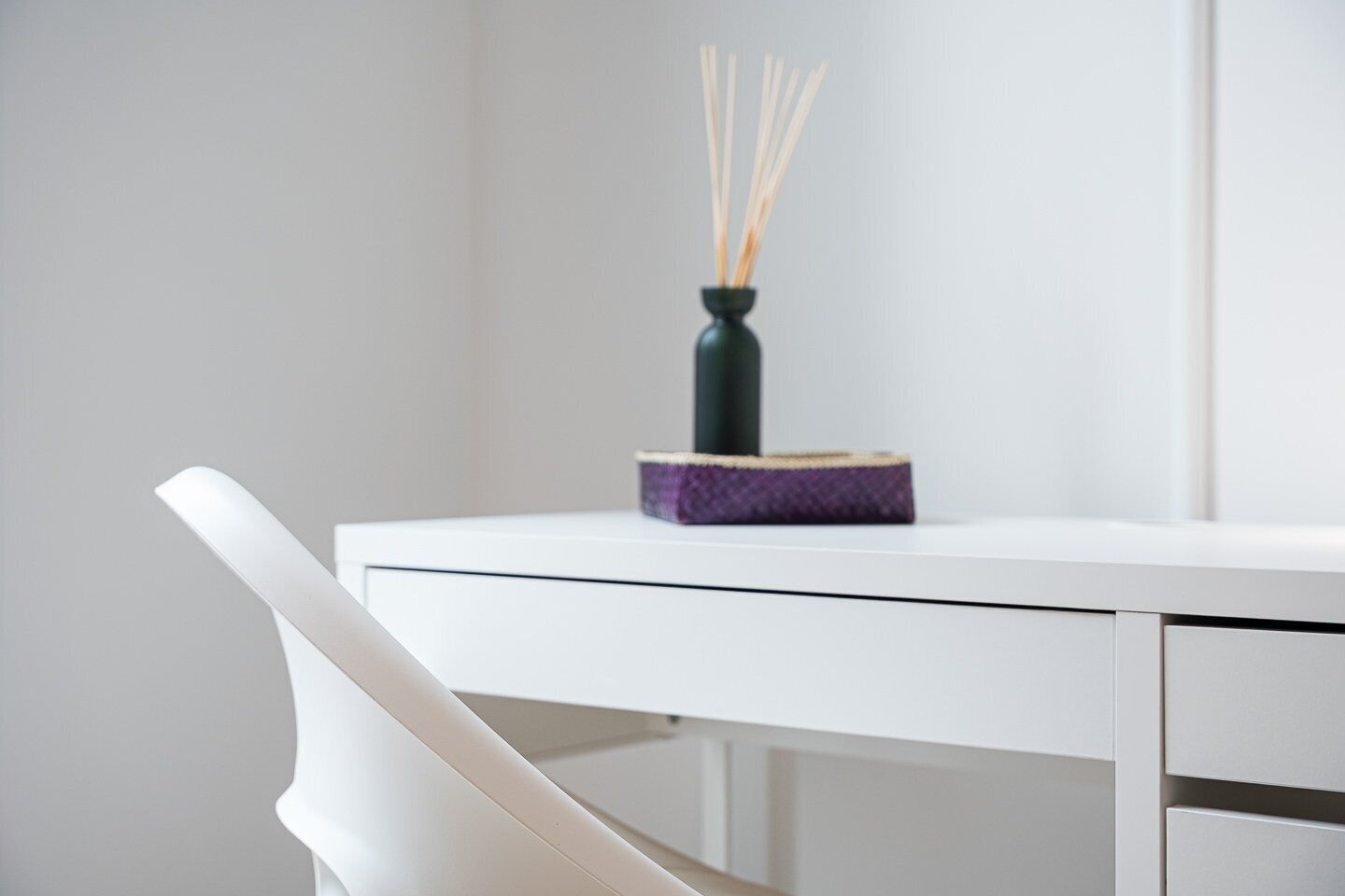 A white desk with a purple book and a green vase with reed sticks.