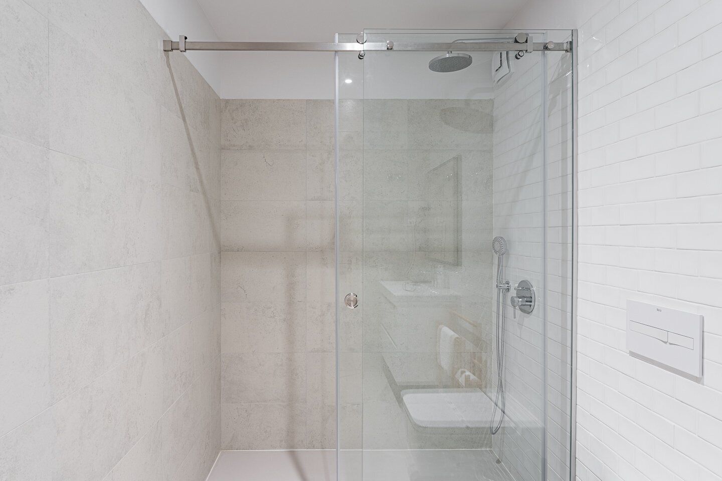 Une cabine de douche fermée par des parois de verre, avec un mur recouvert de carreaux blancs et des toilettes visibles.