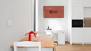 A modern kitchen with a built-in oven, a white dining table with a red gift box, and a flat-screen TV displaying "GUEST READY". - GuestReady – Magnolia (Aveiro)