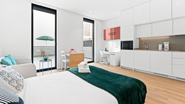 A modern hotel room with a bed, a desk, a chair, a small table with an umbrella, and a kitchen area. - GuestReady – Magnolia (Aveiro)