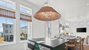 A modern kitchen with a large island, a dining area with a round table, and a view of the beach through large windows. - Beach Side Home, Steps from the Sand. Ocean Edge 14 New Build 2025 (Orange Beach)