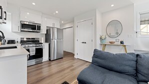 A modern kitchen with white cabinets, stainless steel appliances, and a grey sofa in the living area. - Morden Garden Suite With Parking (Edmonton)