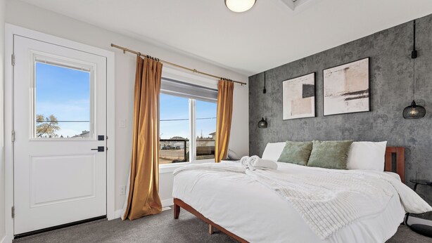 A bedroom with a large bed, a window with curtains, a door, and two framed pictures on the wall. - Morden Garden Suite With Parking (Edmonton)