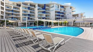 Un hotel moderno con zona de piscina, varios balcones y una terraza de madera con sillones de relax.
