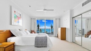 Una habitación de hotel moderna con una cama grande, un ventilador de techo, vistas al océano, una puerta corredera de vidrio y un aparador de madera.