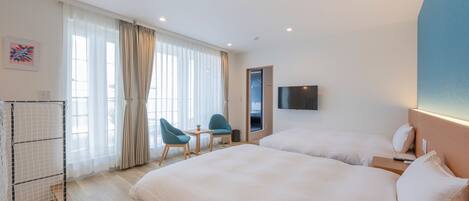 A hotel room with two beds, a TV, a desk with chairs, and a window with curtains.