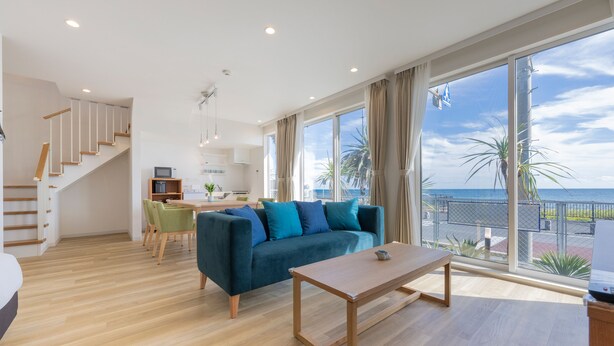 Uma sala de estar ampla com piso de madeira, um sofá azul, uma mesa de centro de madeira e vista para o mar através de grandes janelas.