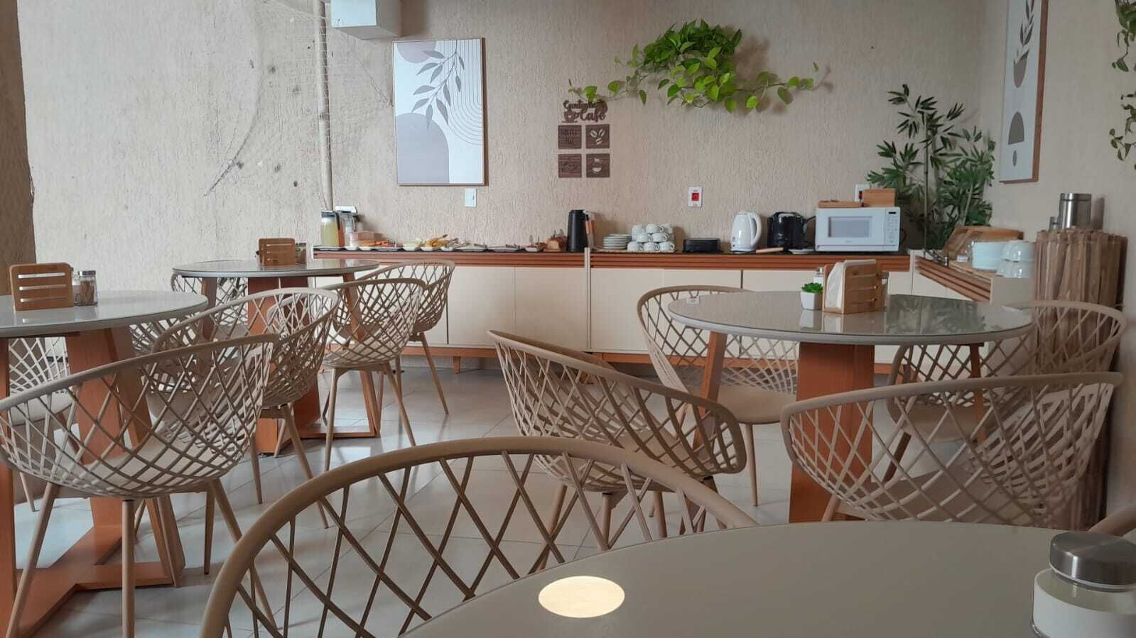 Un café avec des chaises et des tables en osier, un coin cuisine équipé d’appareils électroménagers et agrémenté de plantes décoratives.