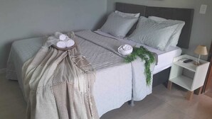 A neatly made bed with a grey headboard, a bedside table with a lamp, and a small shelf. - Canoa dos Anjos Suites (Arraial do Cabo)