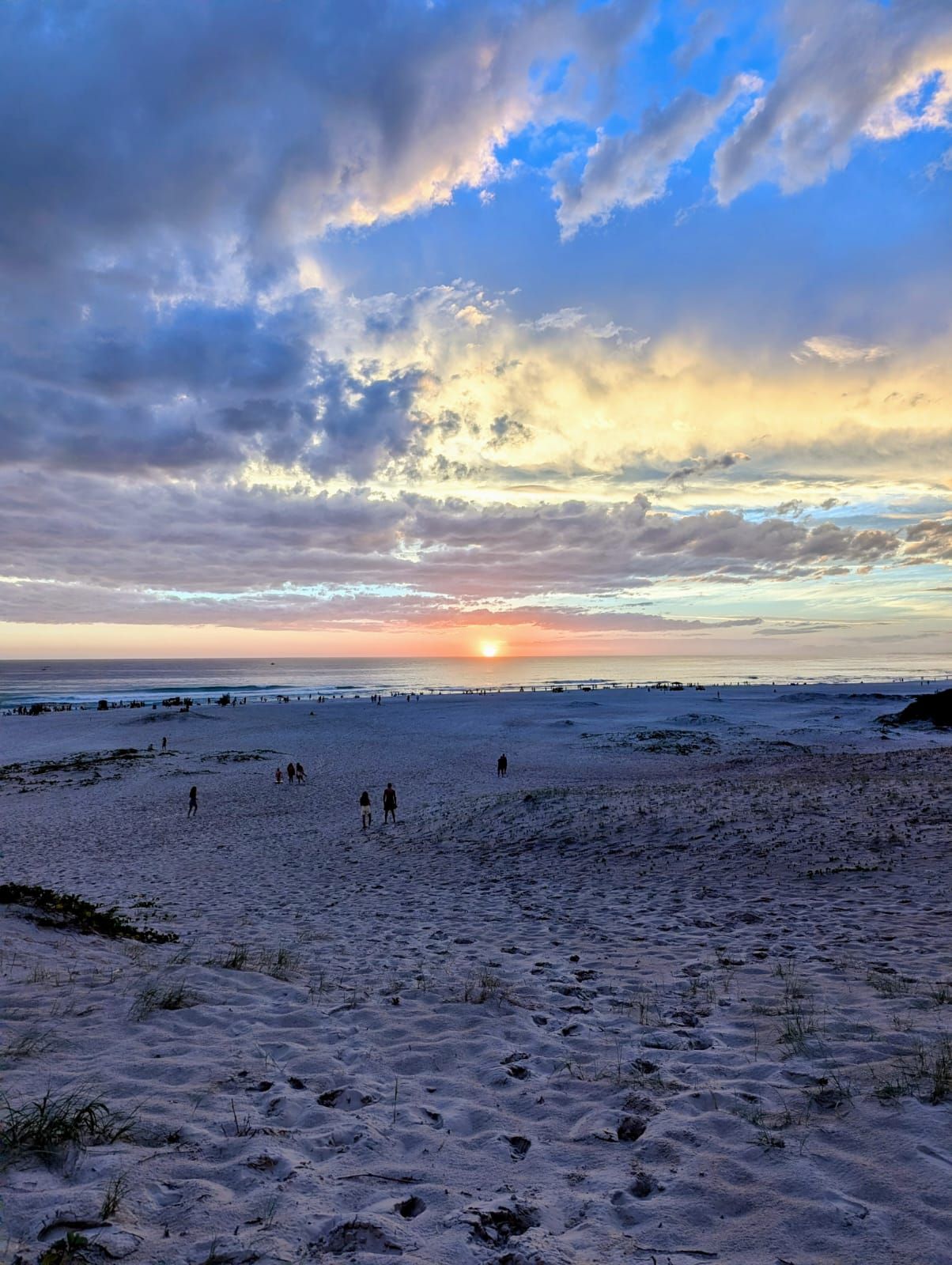 Ein Strand mit Spaziergängern, ein Sonnenuntergang im Hintergrund und ein bewölkter Himmel.