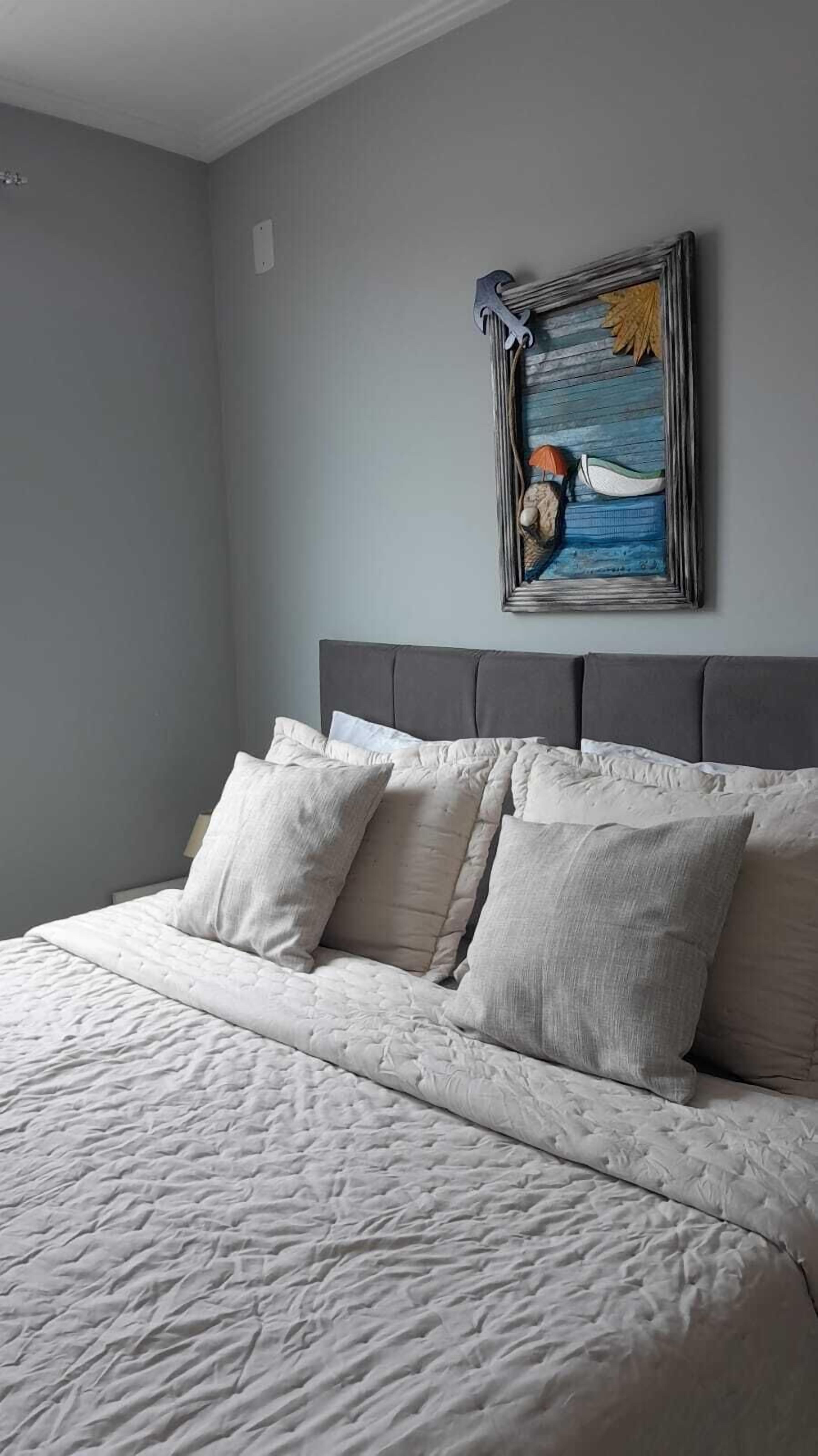 A neatly made bed with a grey headboard and multiple pillows, a framed artwork above it, and a plain grey wall.