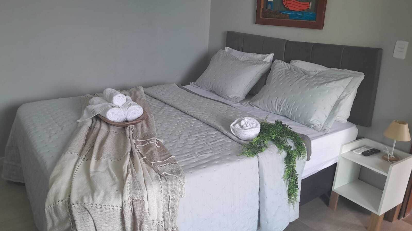 Una cama bien hecha con edredón gris, almohadas y una planta decorativa. Hay una mesita de noche con una lámpara y un teléfono.