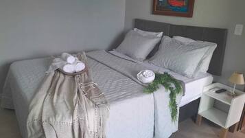 Una cama bien hecha con edredón gris, almohadas y una planta decorativa. Hay una mesita de noche con una lámpara y un teléfono.