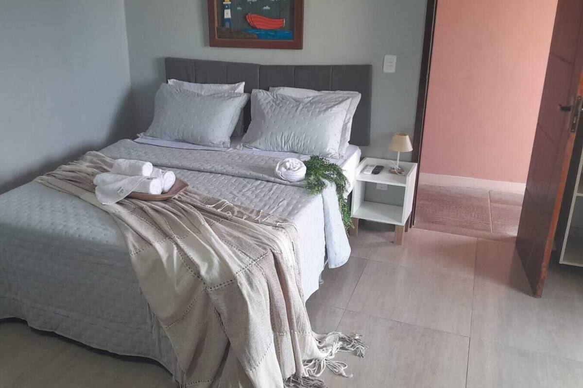 Un lit bien fait, avec une couette blanche et un plaid beige, une table de chevet avec une lampe, et un tableau encadré au mur.