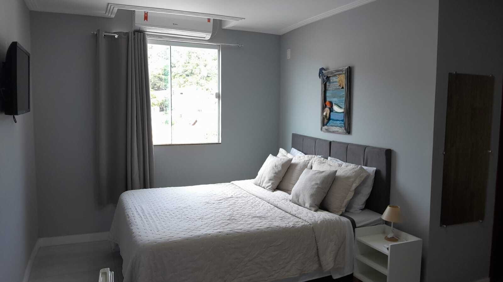 A bedroom with a bed, a nightstand, a lamp, a window with curtains, and a wall-mounted TV.