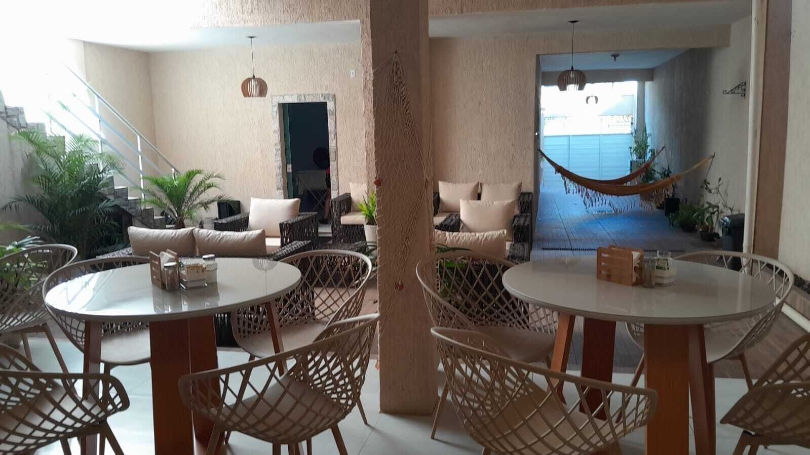 A spacious indoor area with round tables and wicker chairs, a hammock, and a view of the sea through an open doorway.