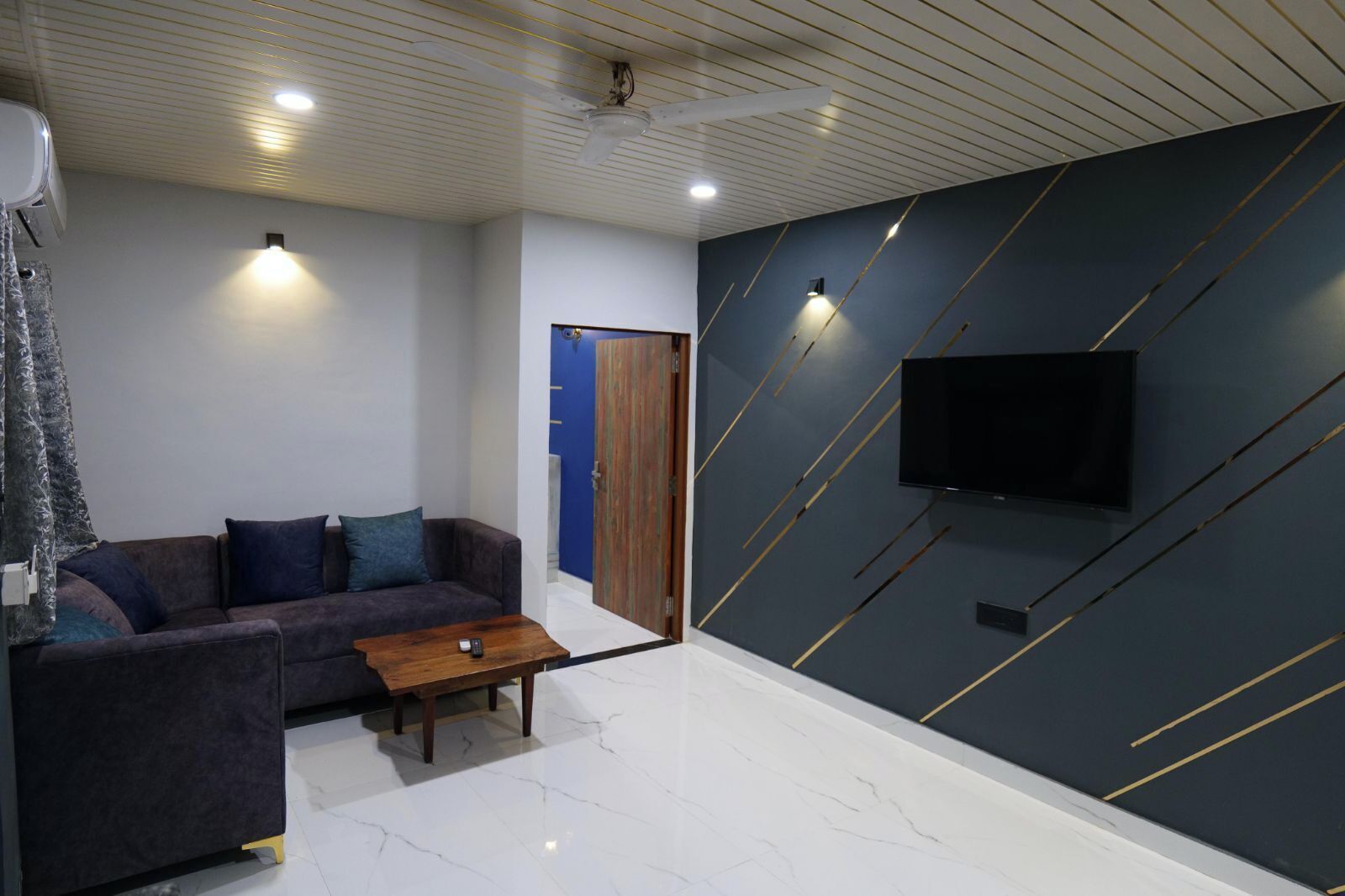 A modern living room with a dark-colored sofa, a wooden coffee table, a flat-screen TV mounted on a dark wall with gold lines, and a ceiling with recessed lighting.