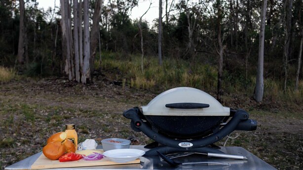 Una parrilla Weber portátil con una tabla de cortar, una botella de salsa y algunos alimentos sobre una superficie metálica al aire libre.