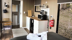 Habitación compacta con cama, un escritorio pequeño, una silla y una kitchenette con microondas, cafetera y extintor.