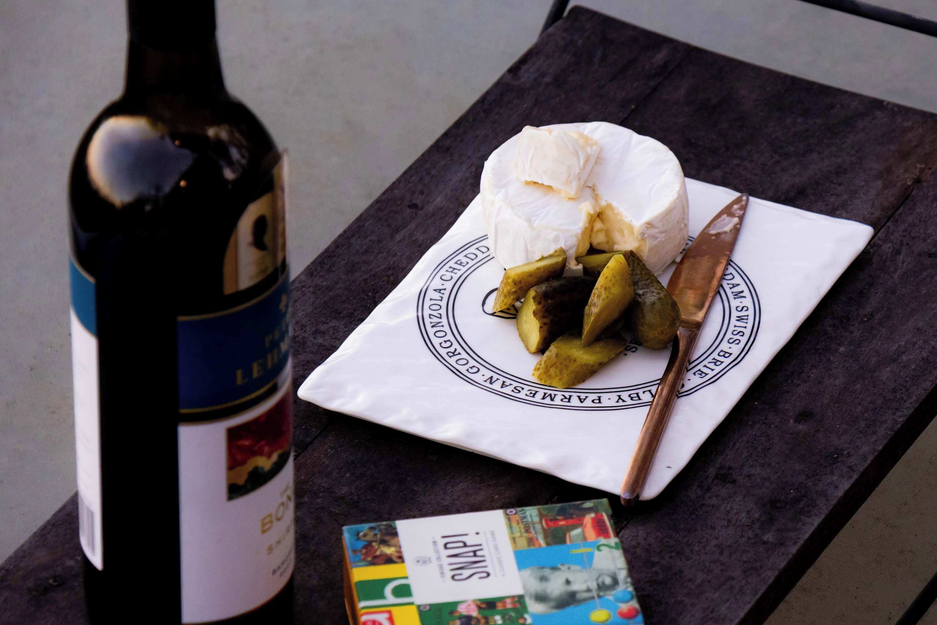 A bottle of wine, a plate with cheese and pickles, and a book on a table.