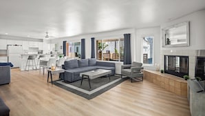 A modern living room with a grey sofa, coffee table, and armchairs. There is a kitchen area with white cabinets and a dining table with chairs. A fireplace is visible on the right side. - Mission Beach 10BR · Steps to Bay & Ocean · Water Views · Firetable (San Diego)