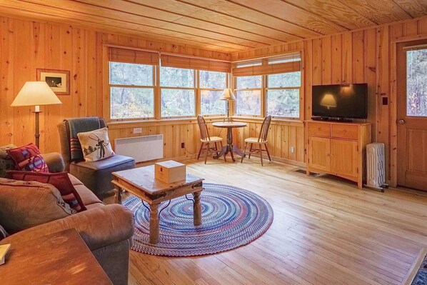 A wooden-paneled room with a sofa, chairs, a table, a TV, and large windows.
