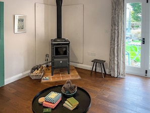 A room with a wood-burning stove, a small stool, a table with books and pinecones, and a door leading to an outdoor area. - The Cowshed - UK50803 (Cratfield, near Laxfield)
