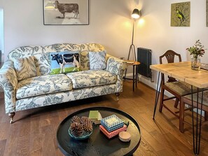 A living room with a patterned sofa, a coffee table with decorative items, a dining table with chairs, and wall art. - The Cowshed - UK50803 (Cratfield, near Laxfield)