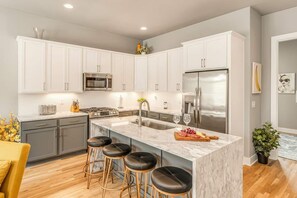 A modern kitchen with white cabinets, a stainless steel refrigerator, a marble island with bar stools, and a central island sink. - Summer rooftop lounge, fire pit, grill near Broadway and Nissan Stadium (Nashville)