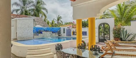Zona de piscina exterior con mesa y sillas de vidrio, rodeada de palmeras y un muro de piedra.