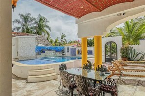 Zona de piscina exterior con mesa y sillas de vidrio, rodeada de palmeras y un muro de piedra.