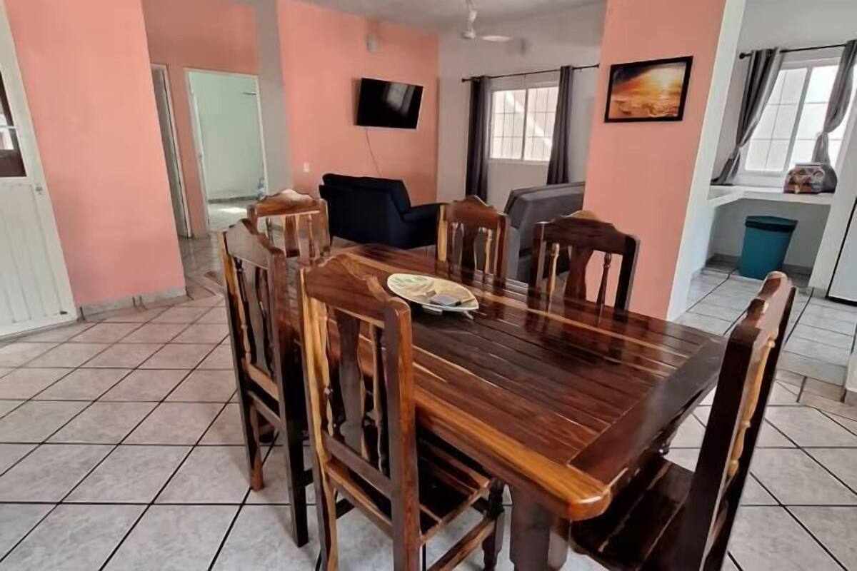 A dining area with a wooden table and chairs, a television mounted on the wall, and a window with curtains.