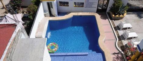 Piscina com deck de azulejos azuis, cercada por um muro branco com portão e plantas em vasos.