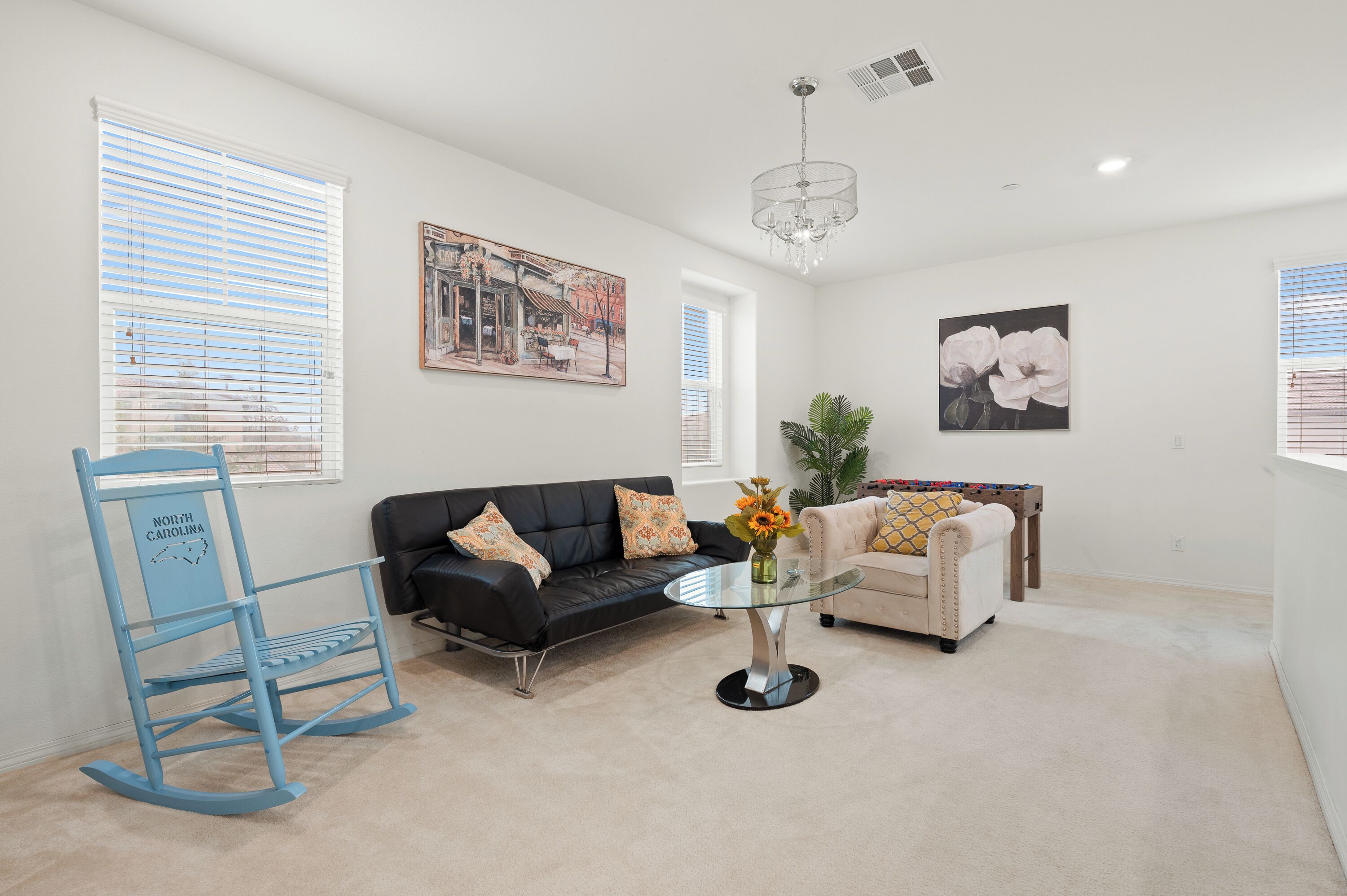 A living room with a black leather sofa, a beige armchair, a glass coffee table, a blue rocking chair, and a chandelier.