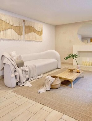 Ein minimalistisches Wohnzimmer mit einem weißen Sofa, einem Holztisch, einer Pflanze und einer Wanddekoration aus Makramee.
