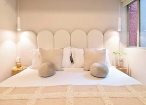 Ein Schlafzimmer mit einem großen Bett, einem Kopfteil, Nachttischen, einem Fenster und einer Pflanze.