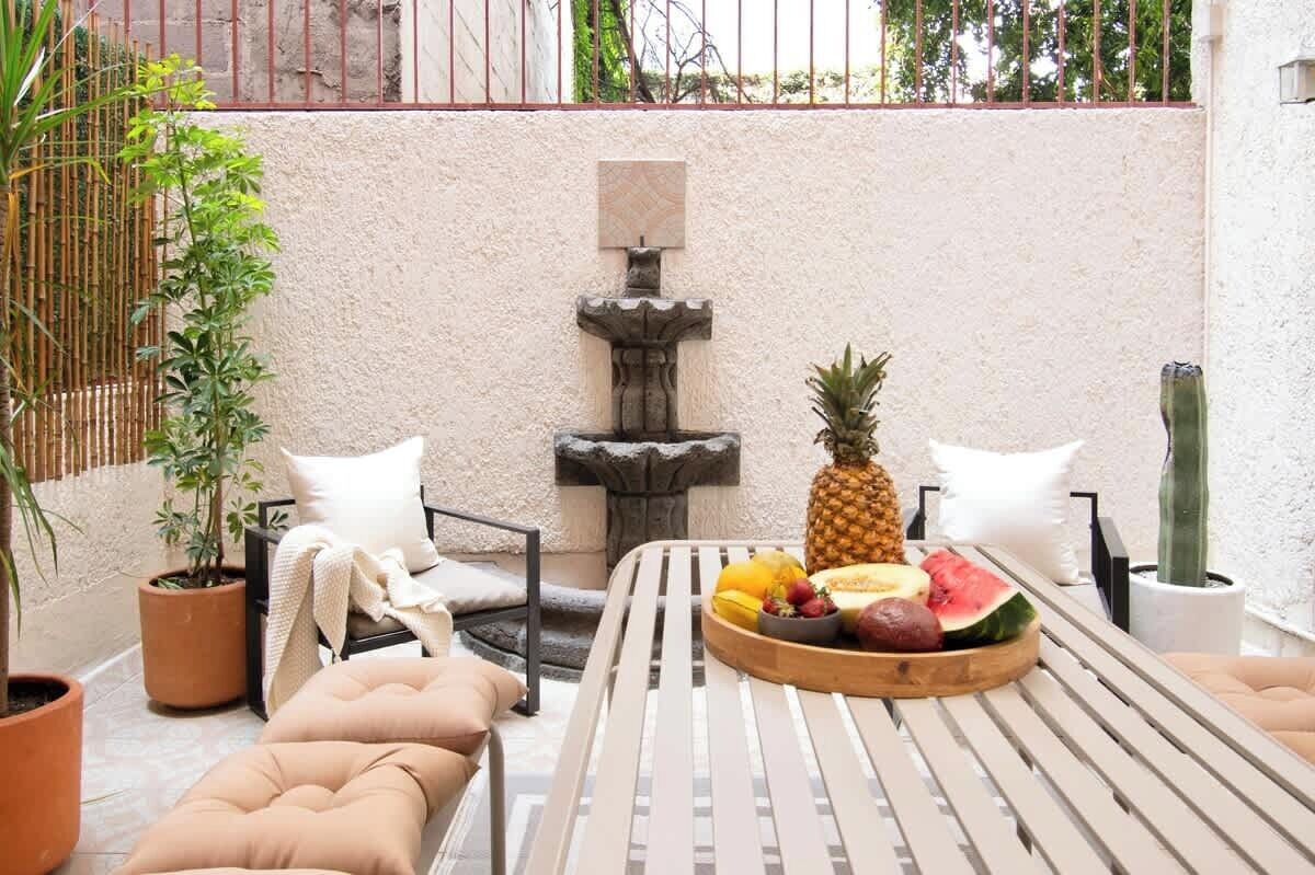 Um pátio com mesa, cadeiras, uma pequena fonte e vasos com plantas.