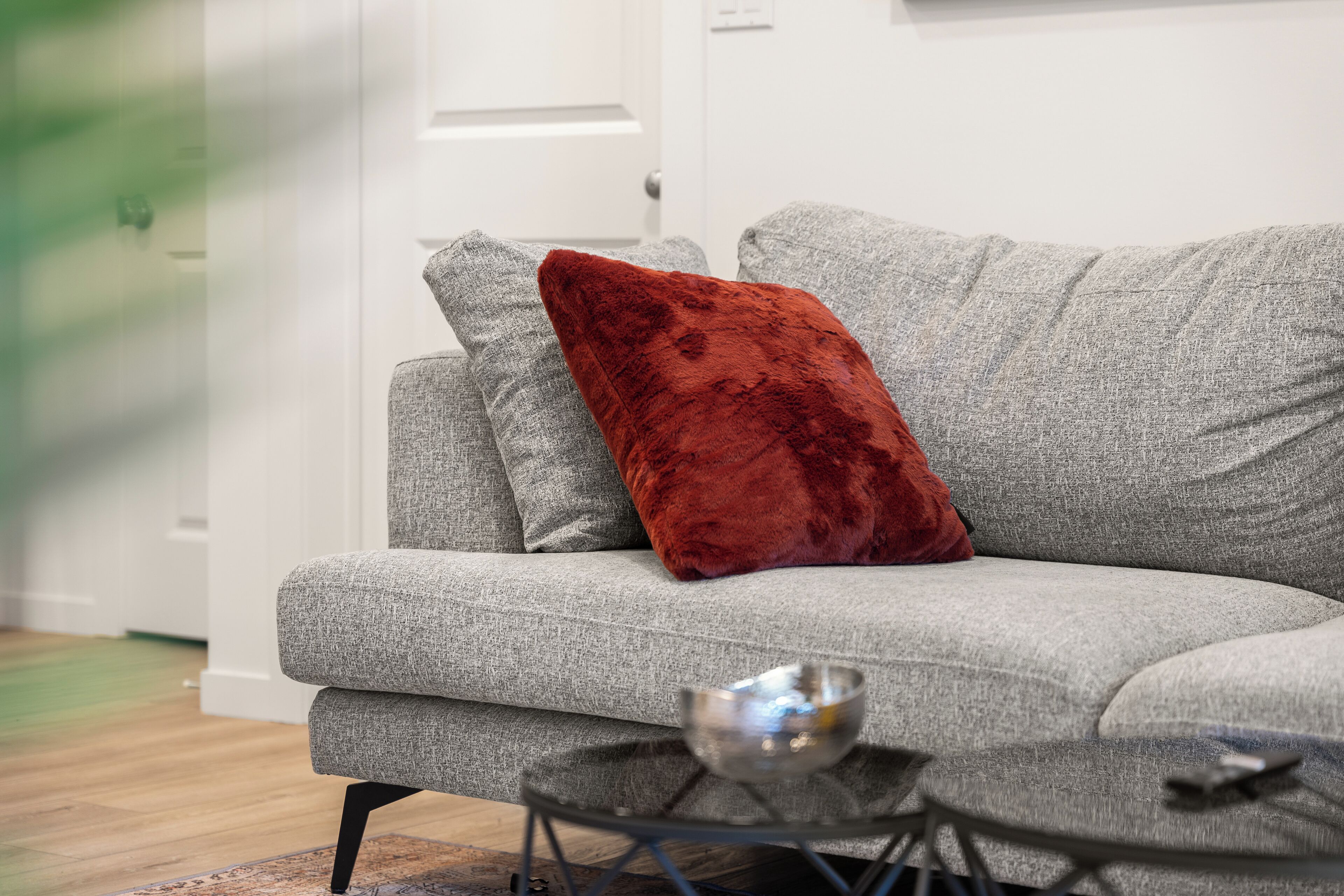Un canapé gris avec un coussin rouge, une petite table ronde surmontée d’un bol en métal et un plancher en bois.