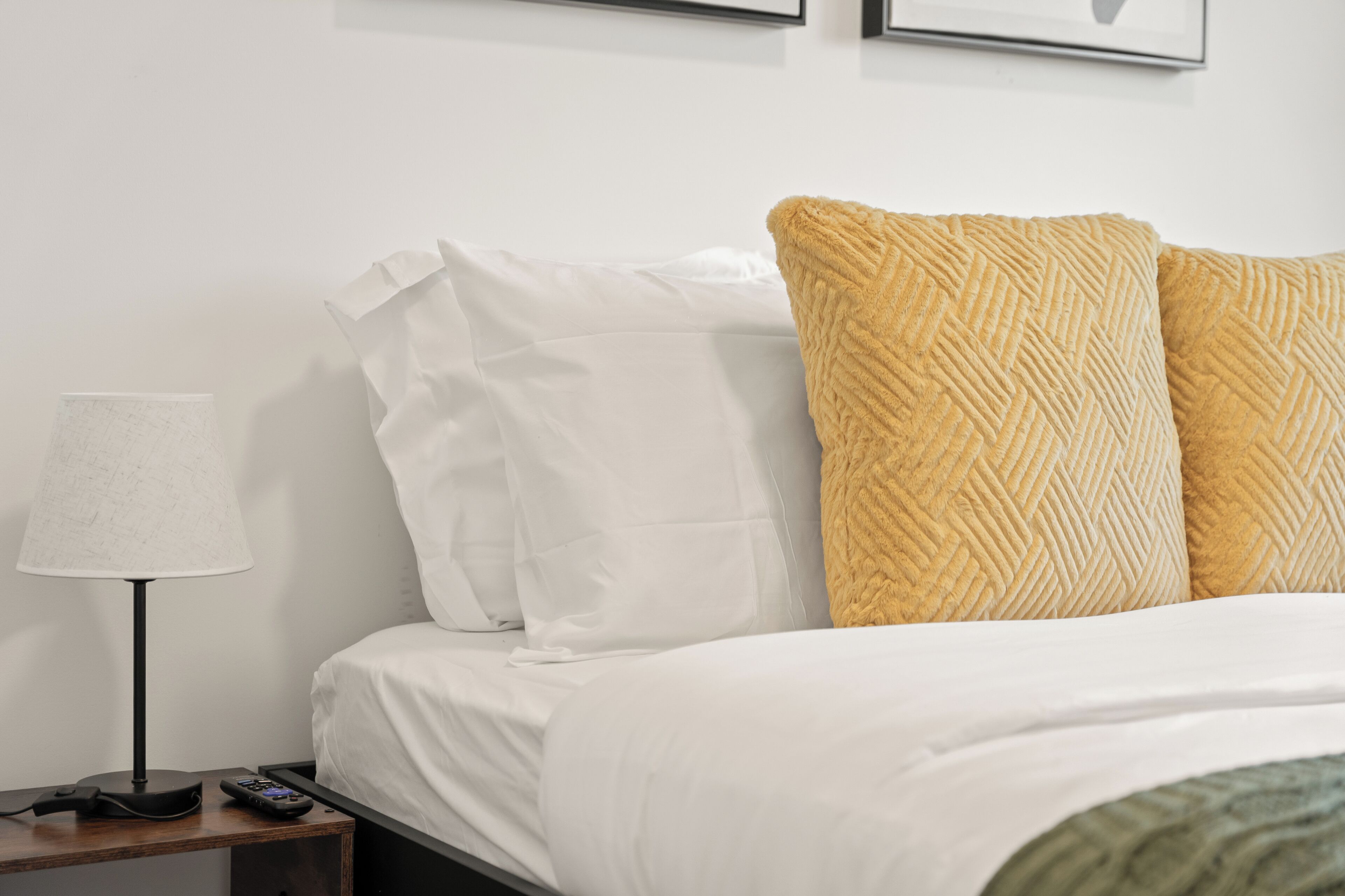 A neatly made bed with white and textured pillows, a lamp on a bedside table, and a remote control.