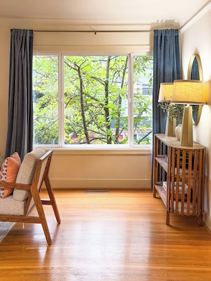 Ein Zimmer mit großem Fenster, Holzboden, einer Couch mit Kissen, einem Beistelltisch mit Lampe und einem kleinen Holzregal.